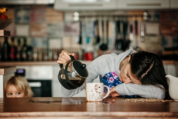 Woman pours hot water from a kettle into a mug on a wooden kitchen counter while a child watches from the left.