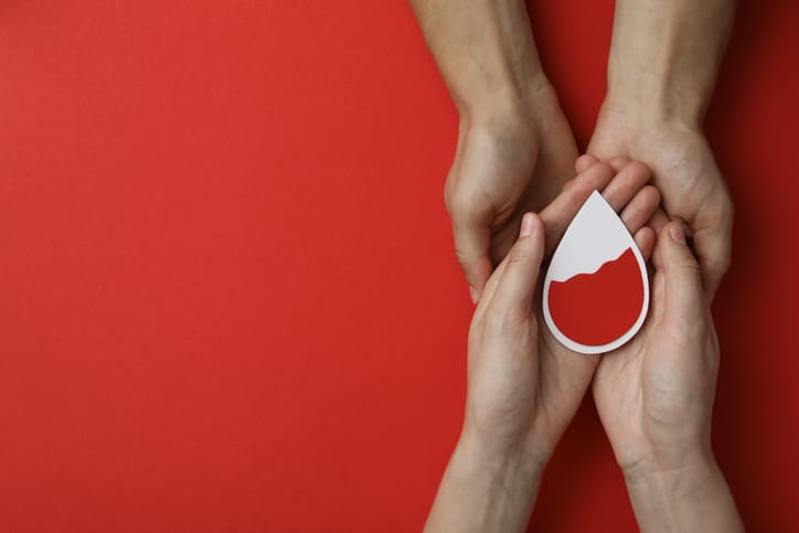 Several hands cradle a white drop-shaped card containing red color, set against a solid red background, symbolizing blood donation.