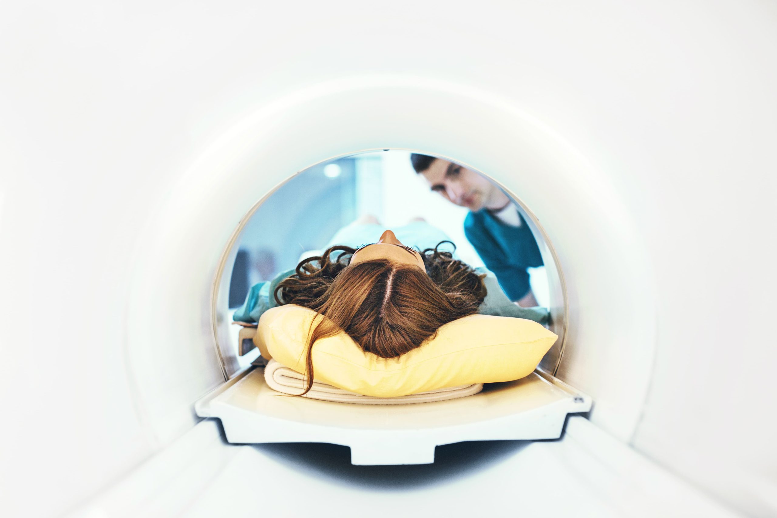 Patient lying in an MRI machine, head on a yellow pillow, with a clinician nearby in scrubs.